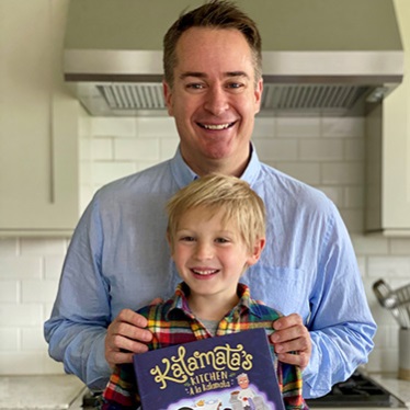 Two individuals standing in a kitchen, one adult and one child, holding a book titled "Kalamatta's Kitchen."