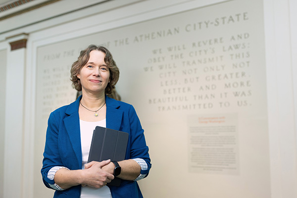 Person in a blue blazer holding a tablet, standing in front of a wall with text about
