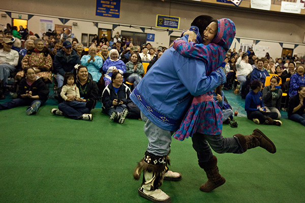 Two people hug joyfully in a gymnasium filled with seated people. The individuals are wearing colorful clothing, and one has a hood. The room appears to host a community event, with banners and applause from onlookers.