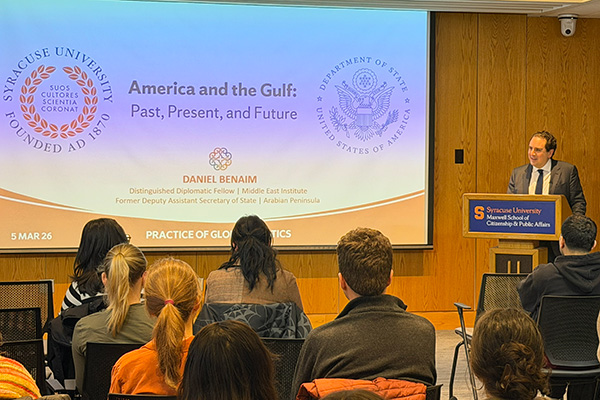 A person giving a presentation at Syracuse University. The screen displays the title