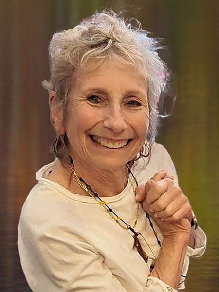 An individual with short grey hair, wearing a white shirt and a necklace, smiles warmly in front of a blurred background.