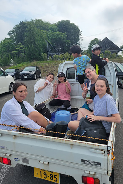 A group of people sitting and standing in the back of a small pickup truck. They appear relaxed and cheerful, some making peace signs. The truck is parked in a lot with several cars in the background. Trees and modern angular buildings are also visible.