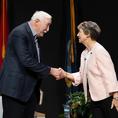 Former U.S. Secretary of State Rex Tillerson and Angela Merkel, former Chancellor of Germany, shaking hands on stage at a public speaking event.