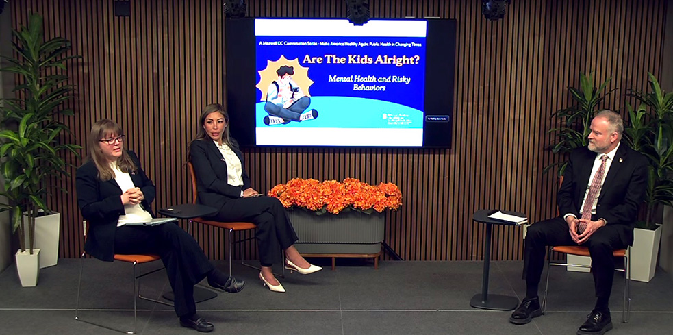A panel discussion setup with three people seated on chairs in a room with wooden panel walls. A screen behind them displays a slide titled