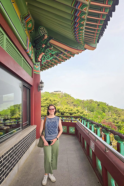 Person standing on a balcony with lush green hills in the background and traditional Korean architecture visible.