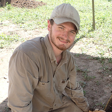 A person in a beige outfit and cap sitting outdoors, smiling at the camera.
