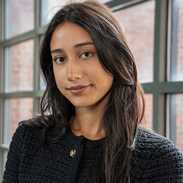 A person wearing a black knit jacket with a small gold pin. They have long dark hair and stands in front of large windows in a brick building.