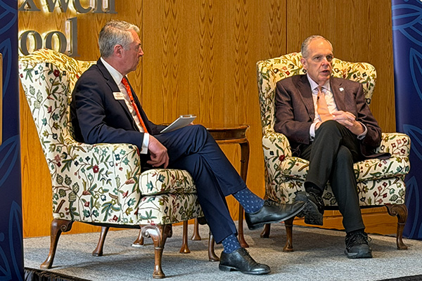Two individuals are seated on stage in patterned armchairs, engaged in a conversation. They are in front of a wooden background that includes the text
