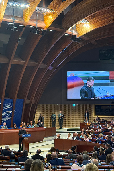 An individual giving a speech at the Council of Europe. The chamber is filled with seated attendees and features a large screen showing the speaker.