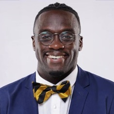A person smiles broadly at the camera, wearing a navy blue blazer, white dress shirt, and black-and-gold striped bow tie. They have short hair and wears glasses, and is photographed against a light gray background.