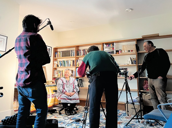 Three people filming a native american woman sitting in a chair