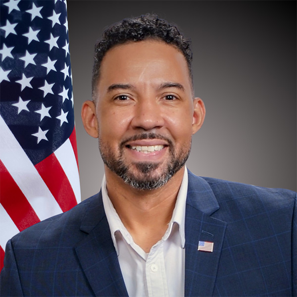 A person smiling wearing a dark suit. There is an American flag in the background.