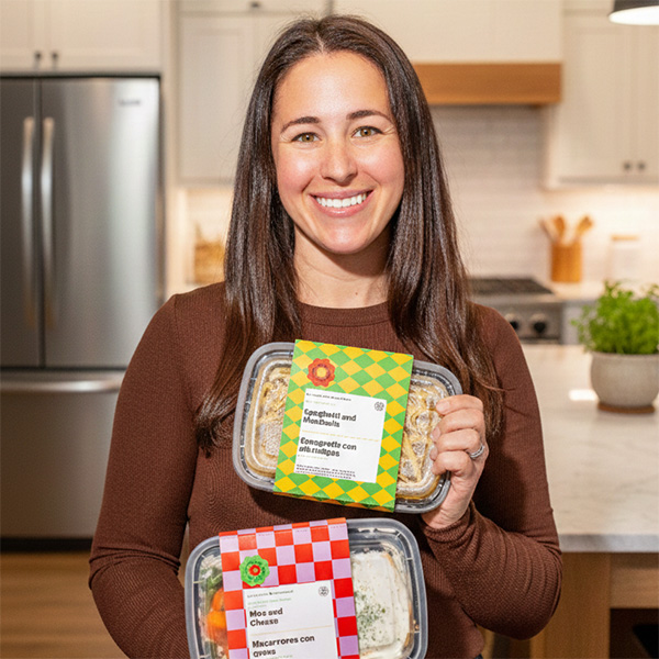 A woman smiles and holds two packaged prepared meals in a kitchen setting. The meal containers are labeled "Spaghetti and Meatballs" and "Mac and Cheese," with bilingual packaging in English and Spanish.